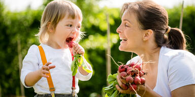 Kleines Kind isst frisch gepflückten roten Rettich, lächelnde Mutter schaut auf sie - draußen im Garten